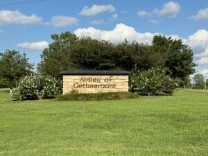 Stone entrance sign reading "Abbey of Gethsemani," flanked by flowering shrubs and trees