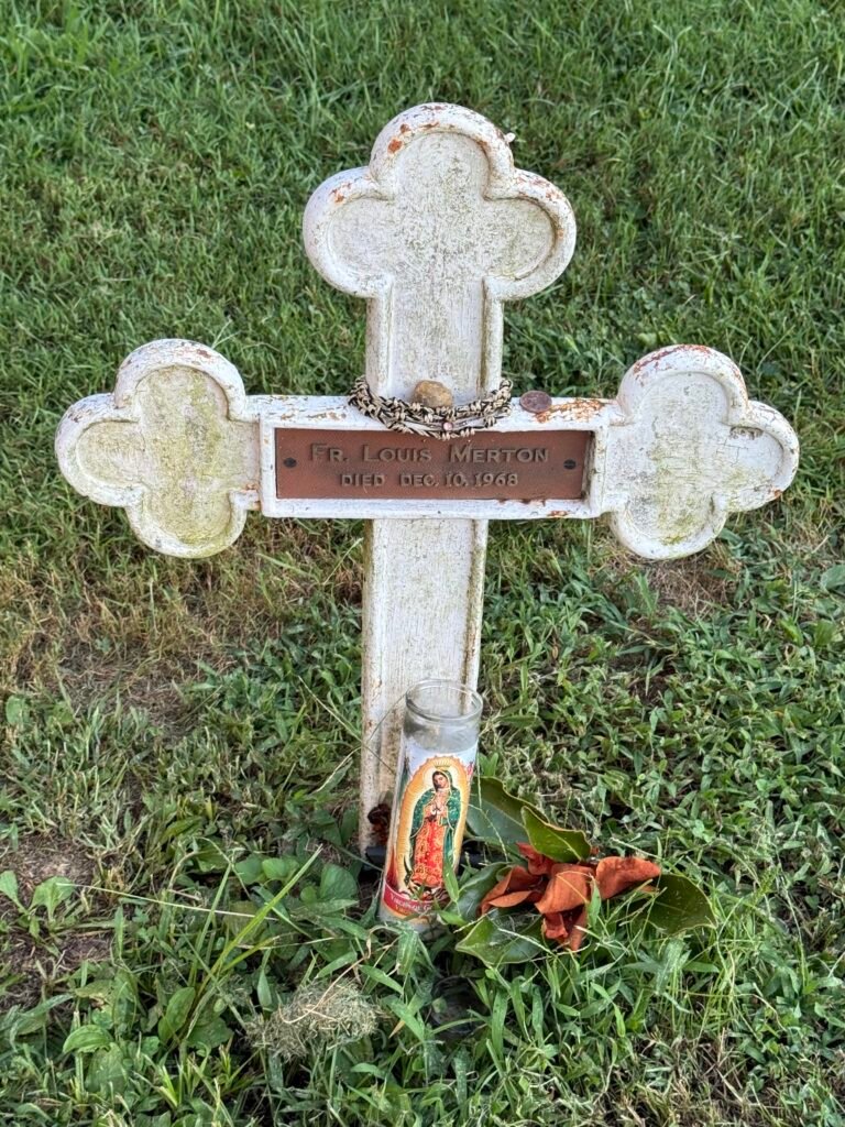 Weathered white cross grave marker reading "Fr. Louis Merton," with a devotional candle and small offerings at its base