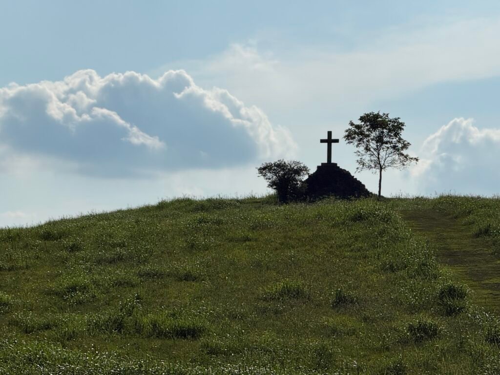 Silhouette of a large cross atop a grassy hilltop against a cloudy sky