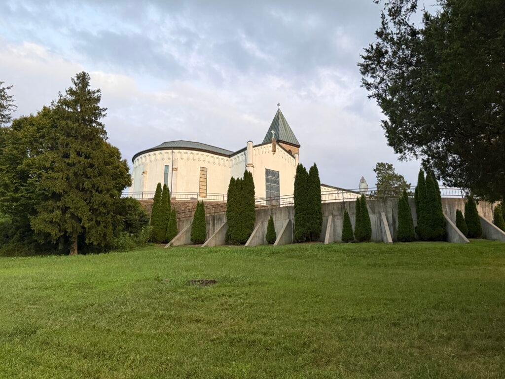 Abbey church exterior at dusk with tiered retaining wall and rows of evergreen trees