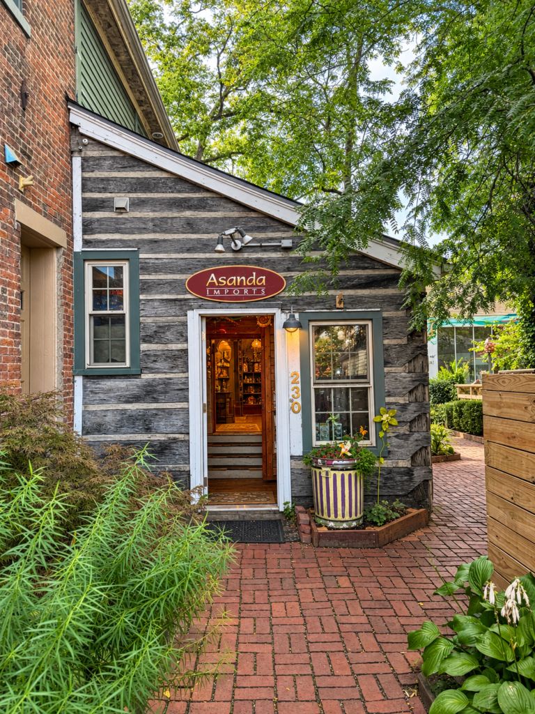 Entrance to Asanda Imports, a small weathered gray clapboard building set back on a brick walkway, with an oval red-and-gold sign above the open door and plantings on either side.