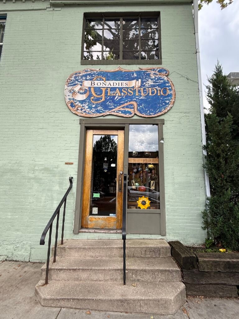 Storefront of Bonadies Glass Studio, pale green painted brick with a weathered blue-and-gold hand-painted sign above a wooden door, concrete steps and iron railings leading up from the sidewalk.
