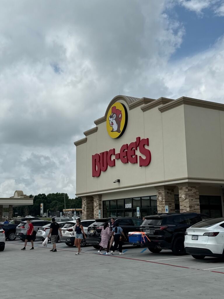Exterior of the Buc-ee's in Huber Heights, Ohio, tan stucco building with the beaver logo and large red BUC-EE'S lettering, shoppers crossing a full parking lot under a partly cloudy sky.