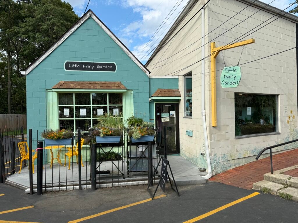 Little Fairy Garden shop, a small teal-painted cinder-block building with a chalkboard sign over the display window, flower boxes and yellow café chairs behind an iron fence, and a hanging sign at the corner entrance.