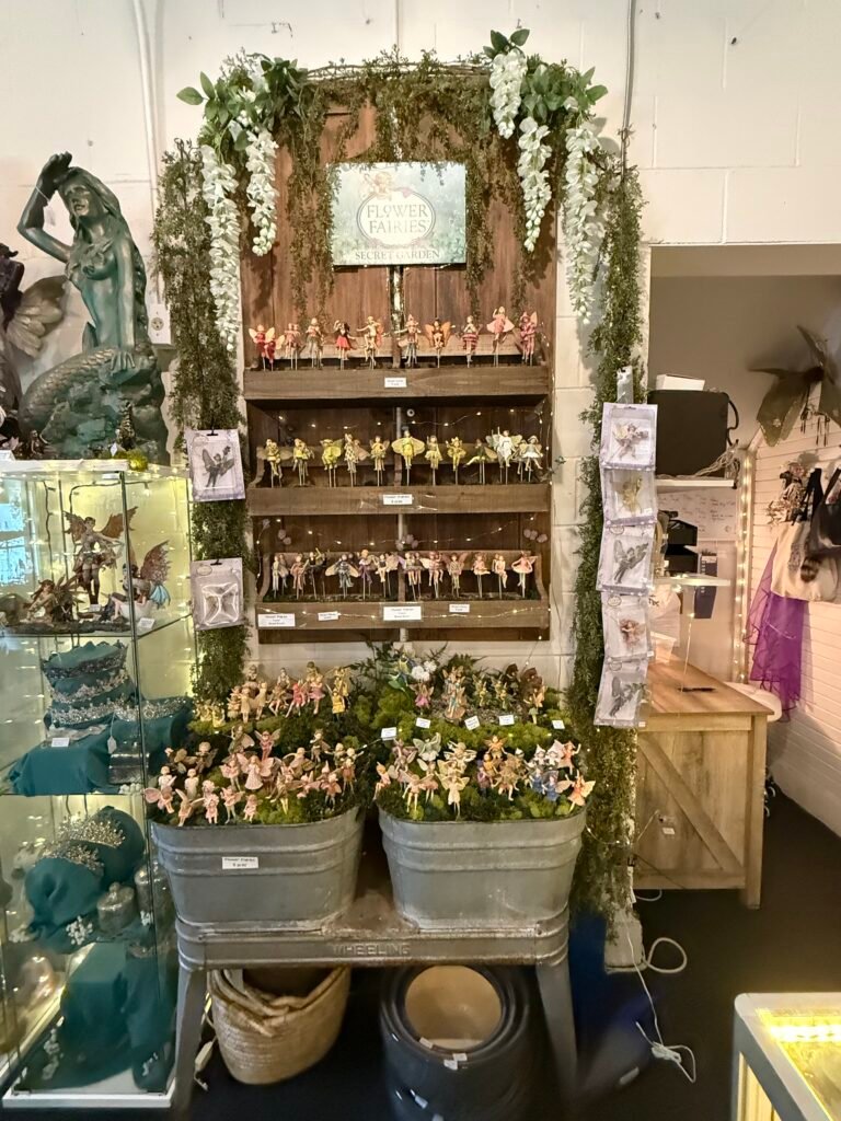 Rustic wood shelving hung with greenery and white blossoms under a Flower Fairies Secret Garden sign, rows of small painted fairy figures on stakes, with two galvanized tubs of loose figurines below.