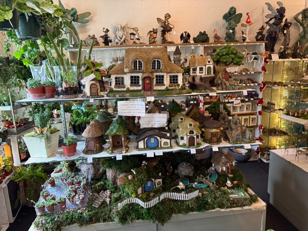 Three glass shelves crowded with miniature fairy houses—thatched cottages, mushroom-capped huts, moss-covered tree stumps—each with a hand-lettered price tag, fairy statues above and a landscaped village display below.