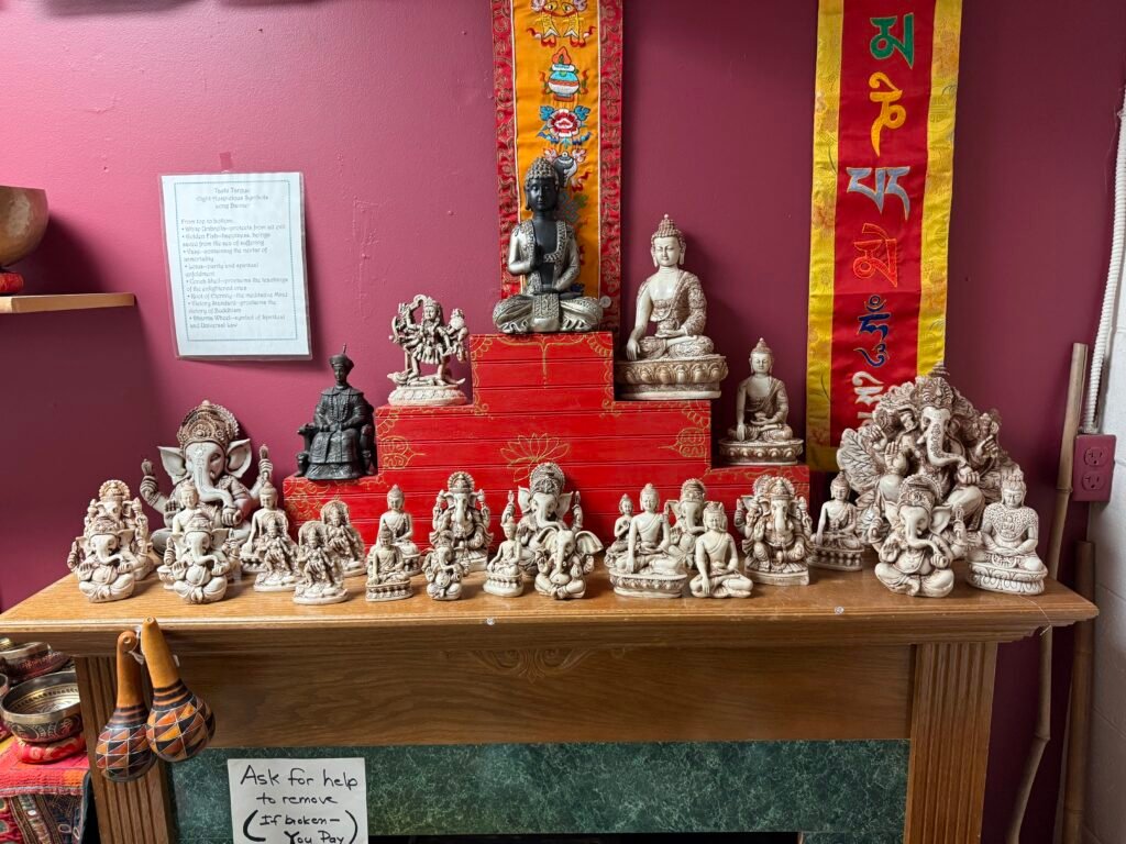 A wooden table against a deep red wall, lined with cream-colored Ganesha and Buddha statues in graduated sizes, red lacquer risers behind them and embroidered Tibetan banners on the wall above.