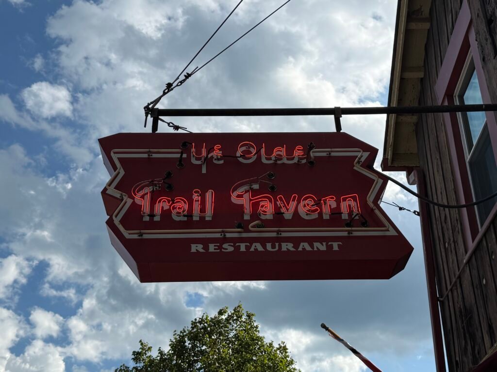 Arrow-shaped red neon sign reading Ye Olde Trail Tavern Restaurant, hung from a bracket against a bright cloudy sky.