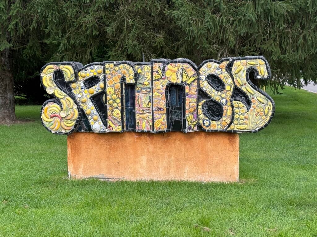 Ceramic mosaic sculpture spelling SPRINGS in yellow tile, mounted on a low orange plinth on a strip of grass between a sidewalk and a road, evergreens behind it.