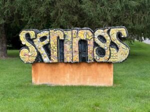 Ceramic mosaic sculpture spelling SPRINGS in yellow tile, mounted on a low orange plinth on a strip of grass between a sidewalk and a road, evergreens behind it.