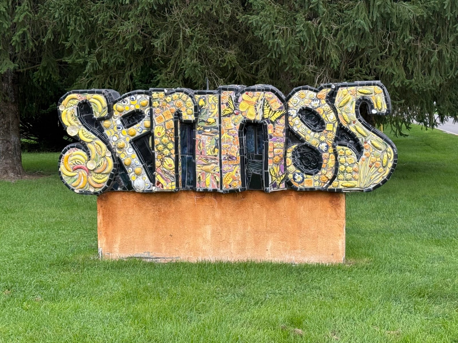 Ceramic mosaic sculpture spelling SPRINGS in yellow tile, mounted on a low orange plinth on a strip of grass between a sidewalk and a road, evergreens behind it.