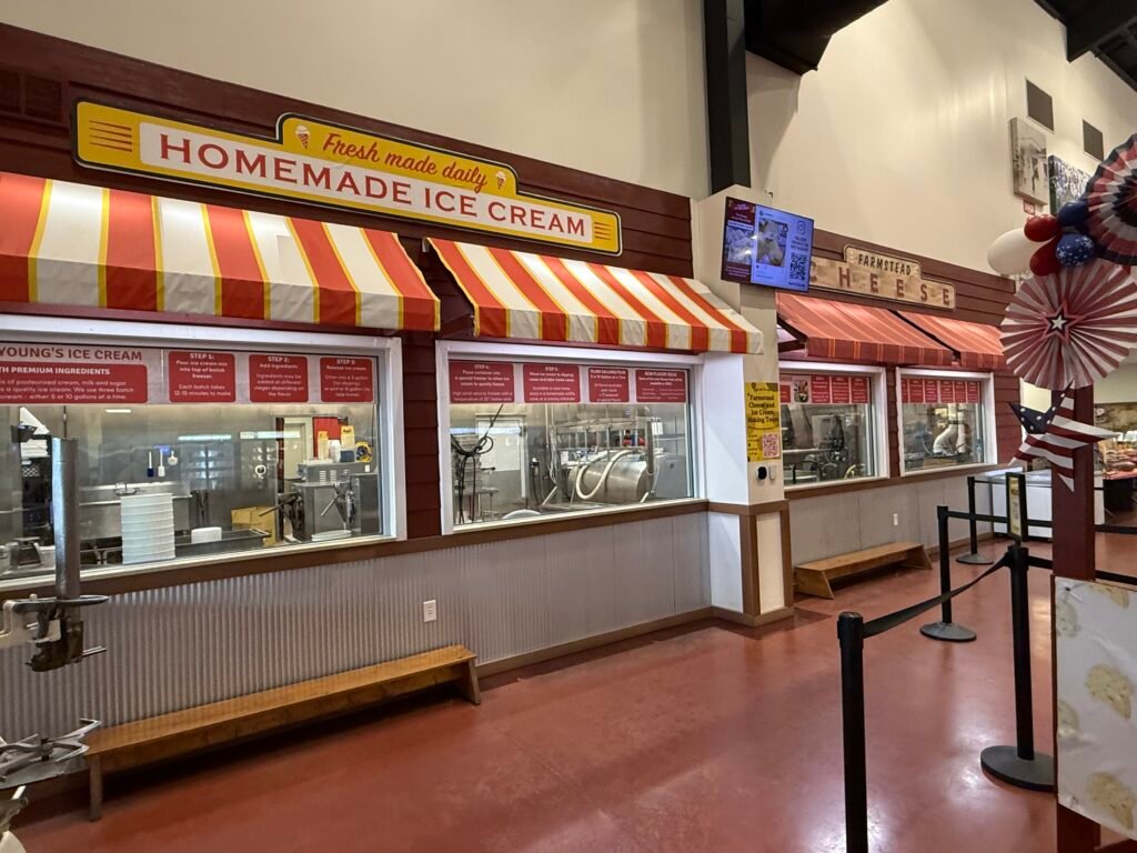 Interior viewing windows at Young's Jersey Dairy, striped awnings over glass panels marked Homemade Ice Cream and Farmstead Cheese, with numbered signs explaining each production step and stainless equipment visible inside.