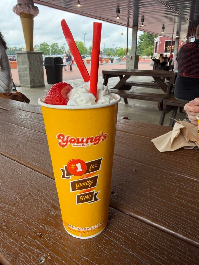 A tall yellow Young's Jersey Dairy cup on a wooden picnic table, filled with a strawberry milkshake topped with whipped cream and a whole strawberry, two red straws in it, the covered patio and parking lot behind.