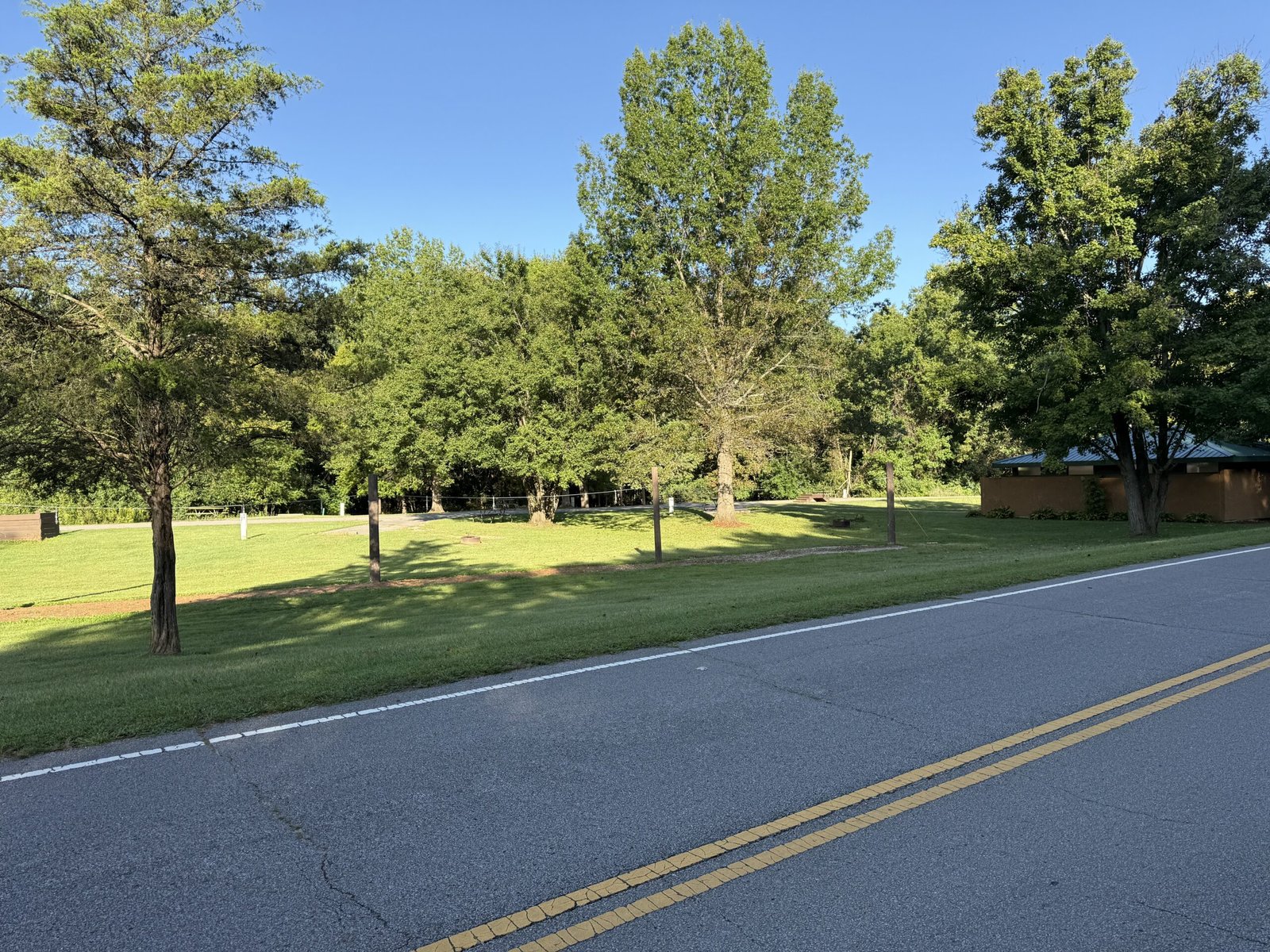 Mown campsites under scattered trees, with brown posts and a strung wire running across the grass in front of a park building