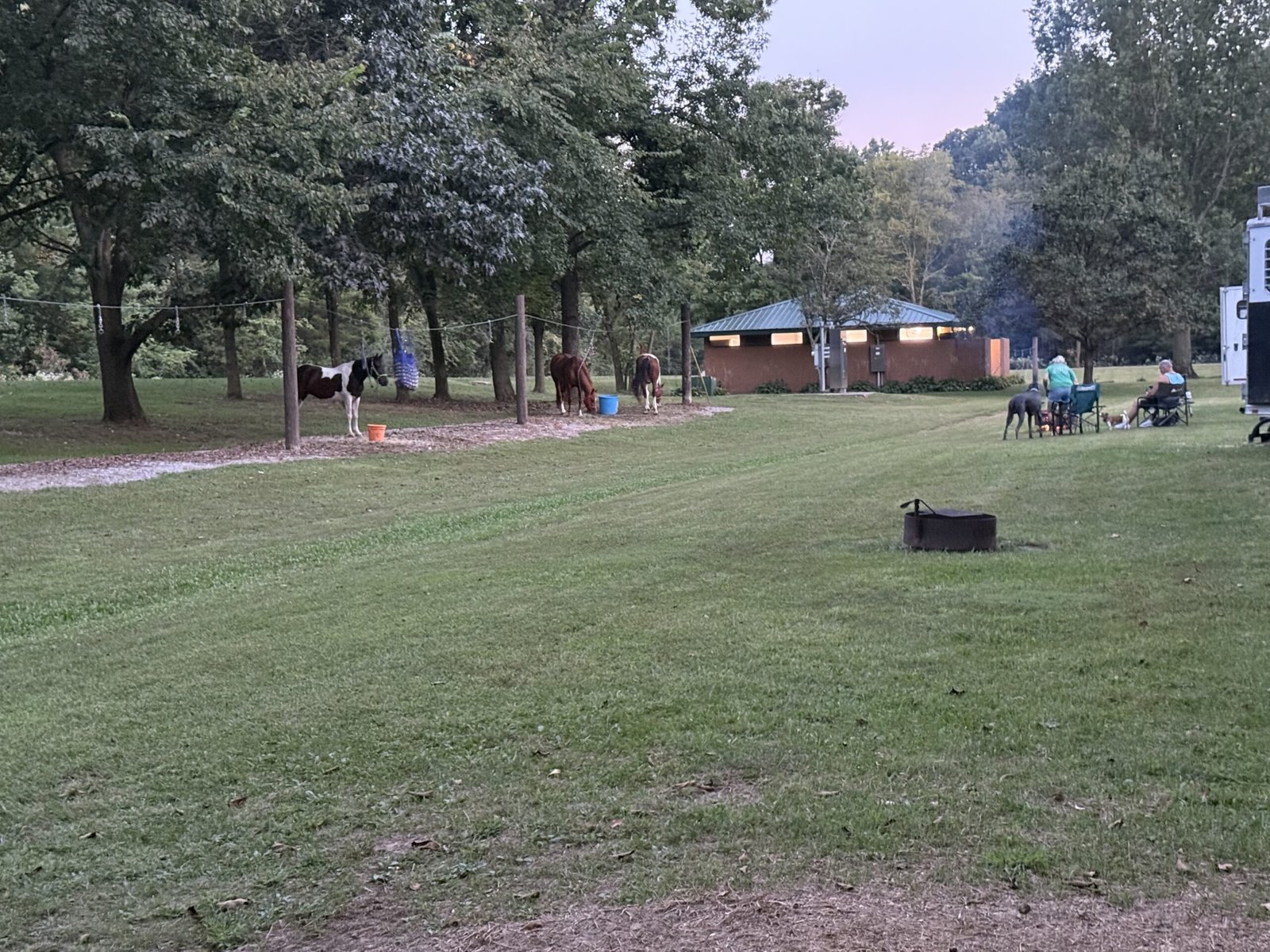The horse camp at dusk, with horses on the picket line, campers seated beside a trailer, and a lit shower house beyond