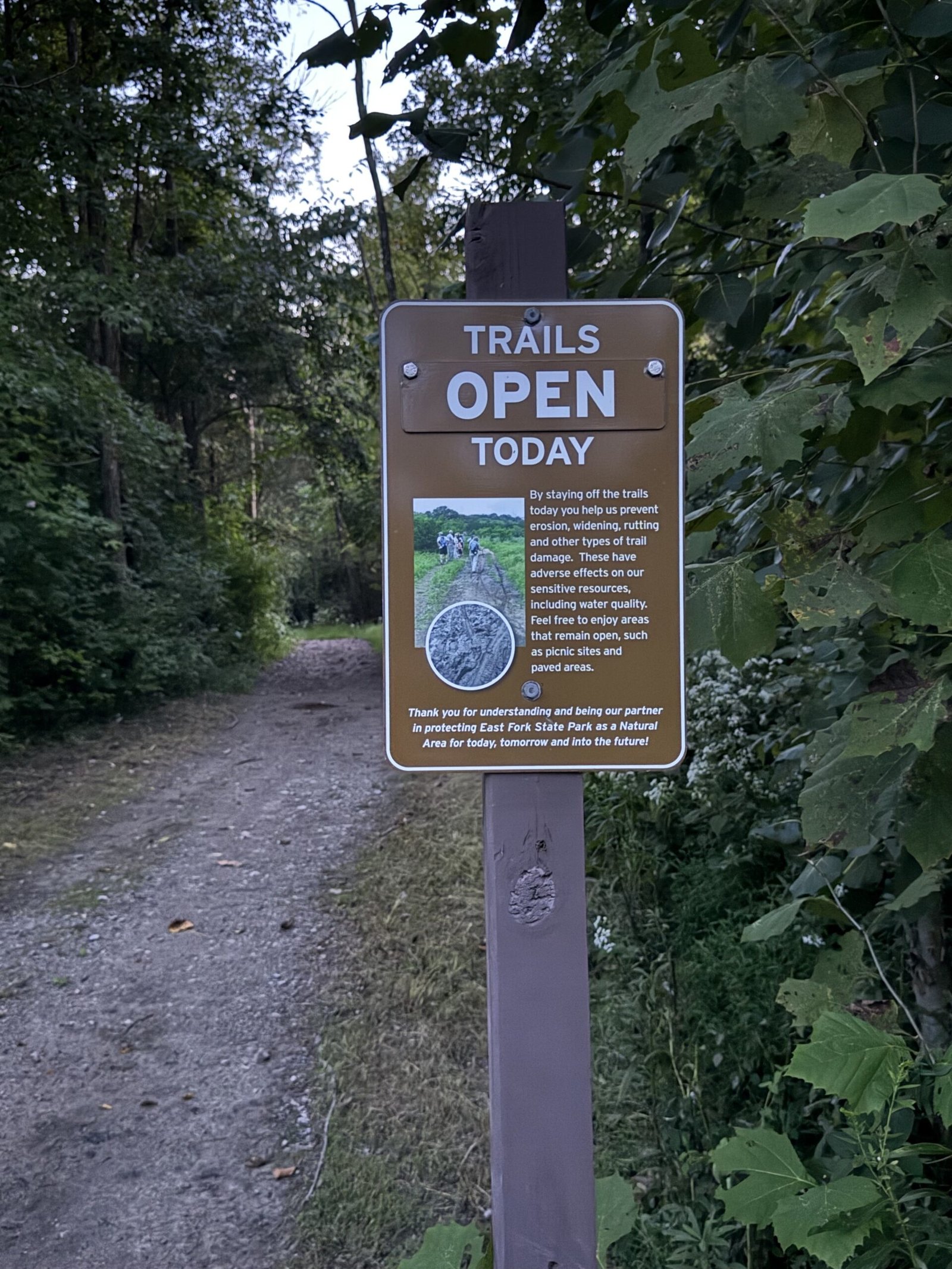 A brown park sign reading Trails Open Today, with text below asking riders to respect closures when conditions would damage the trail