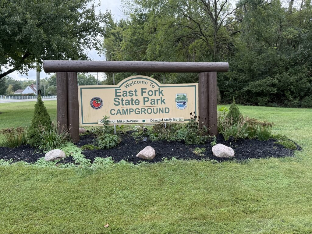 A wooden-framed park sign reading Welcome To East Fork State Park Campground, standing in a landscaped bed of shrubs and stone