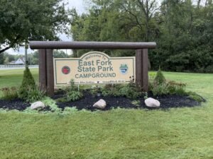 A wooden-framed park sign reading Welcome To East Fork State Park Campground, standing in a landscaped bed of shrubs and stone