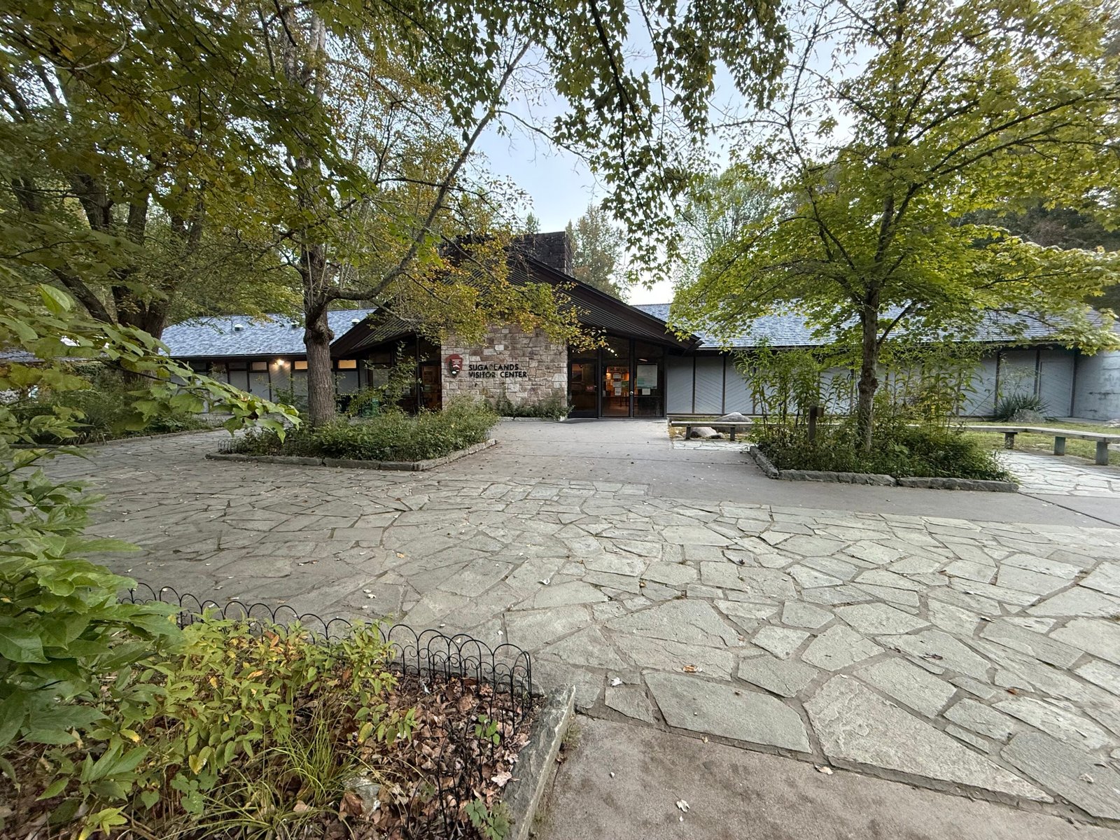 The Sugarlands Visitor Center, a low stone and timber building behind a flagstone plaza and trees in early morning light