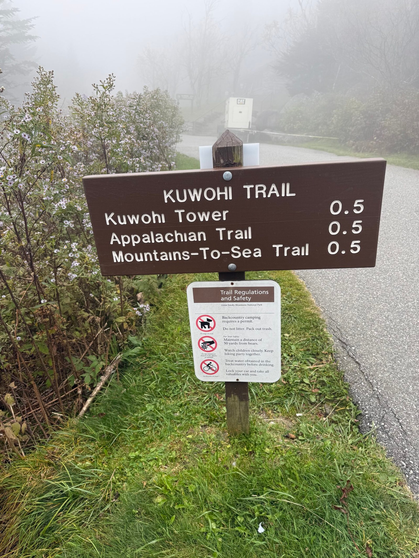 A brown trail sign reading Kuwohi Trail, with the Kuwohi Tower, the Appalachian Trail, and the Mountains-to-Sea Trail each listed at half a mile