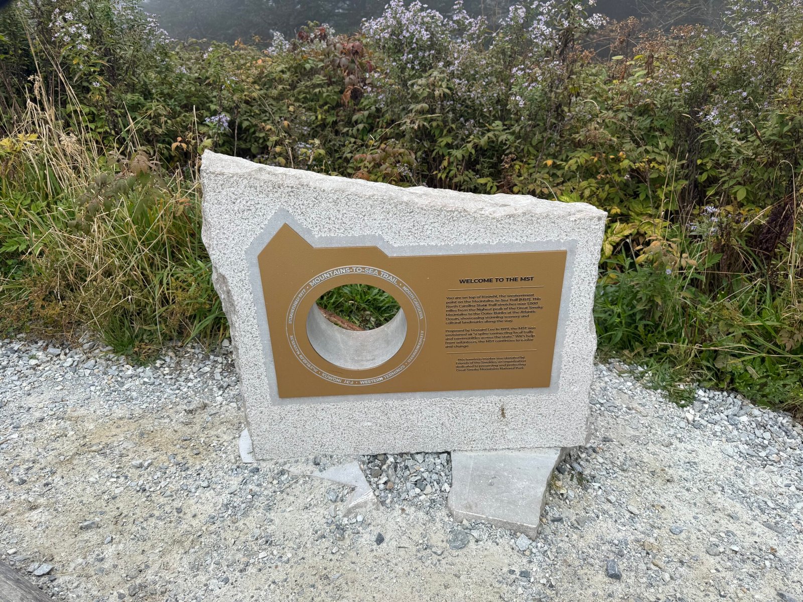 A granite Mountains-to-Sea Trail marker with a bronze plaque reading Welcome to the Mist, set in gravel beside the trail