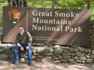 The author sitting on a river-rock wall beneath the brown Great Smoky Mountains National Park entrance sign
